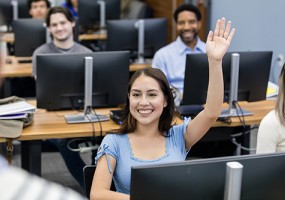 A smiling Latina student in a computer lab raises her hand.