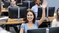 A smiling Latina student in a computer lab raises her hand.